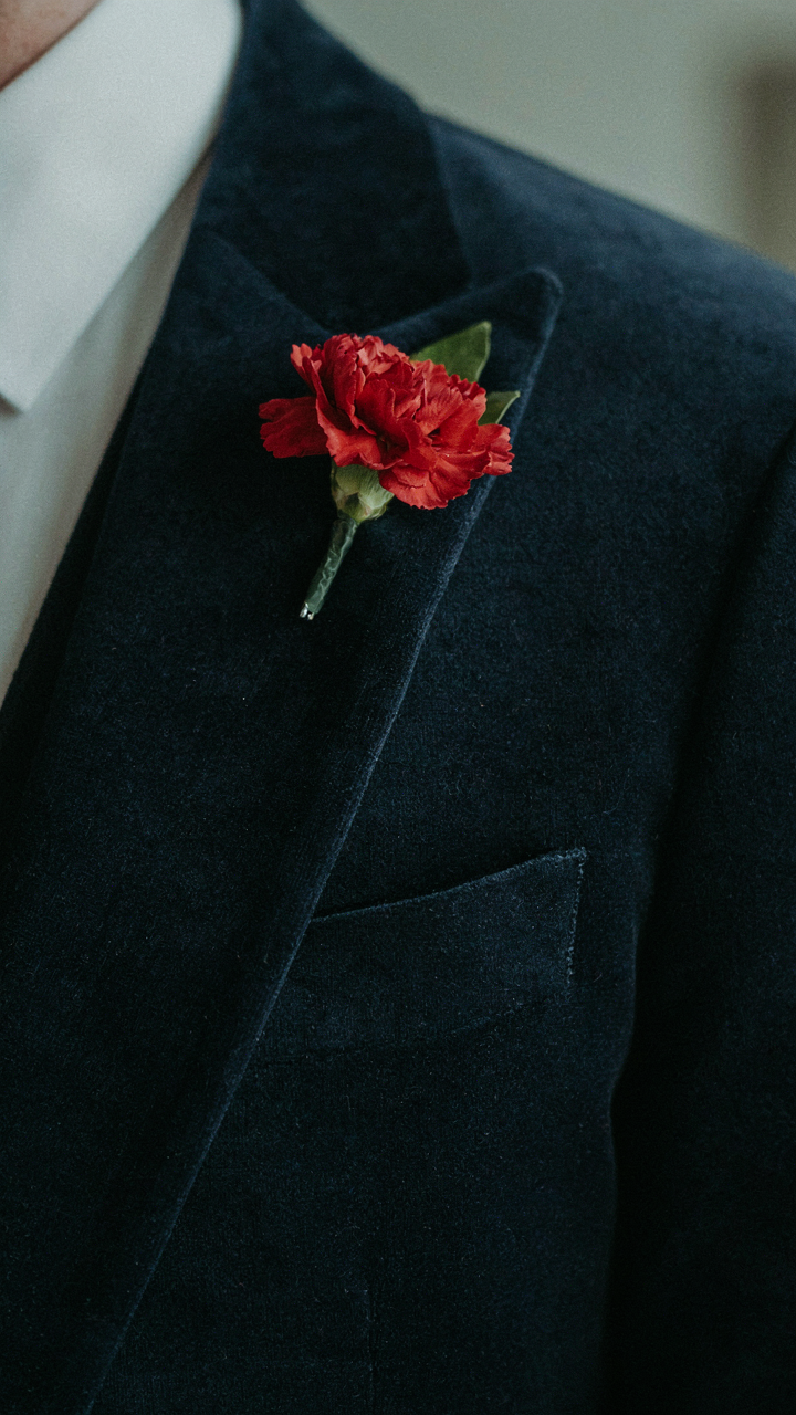 A close-up of a man's velvet jacket lapel with a red boutonniere.