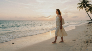 Woman in a linen dress walks on a beach at sunset with a small tote bag.