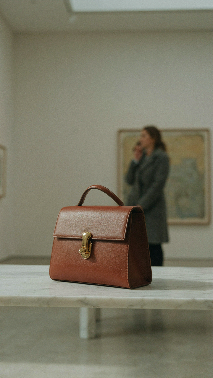 A distinctive structured leather handbag sits on a marble bench in an art gallery.