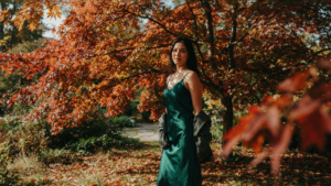 A woman in an emerald october wedding guest dress in a park with autumn leaves.