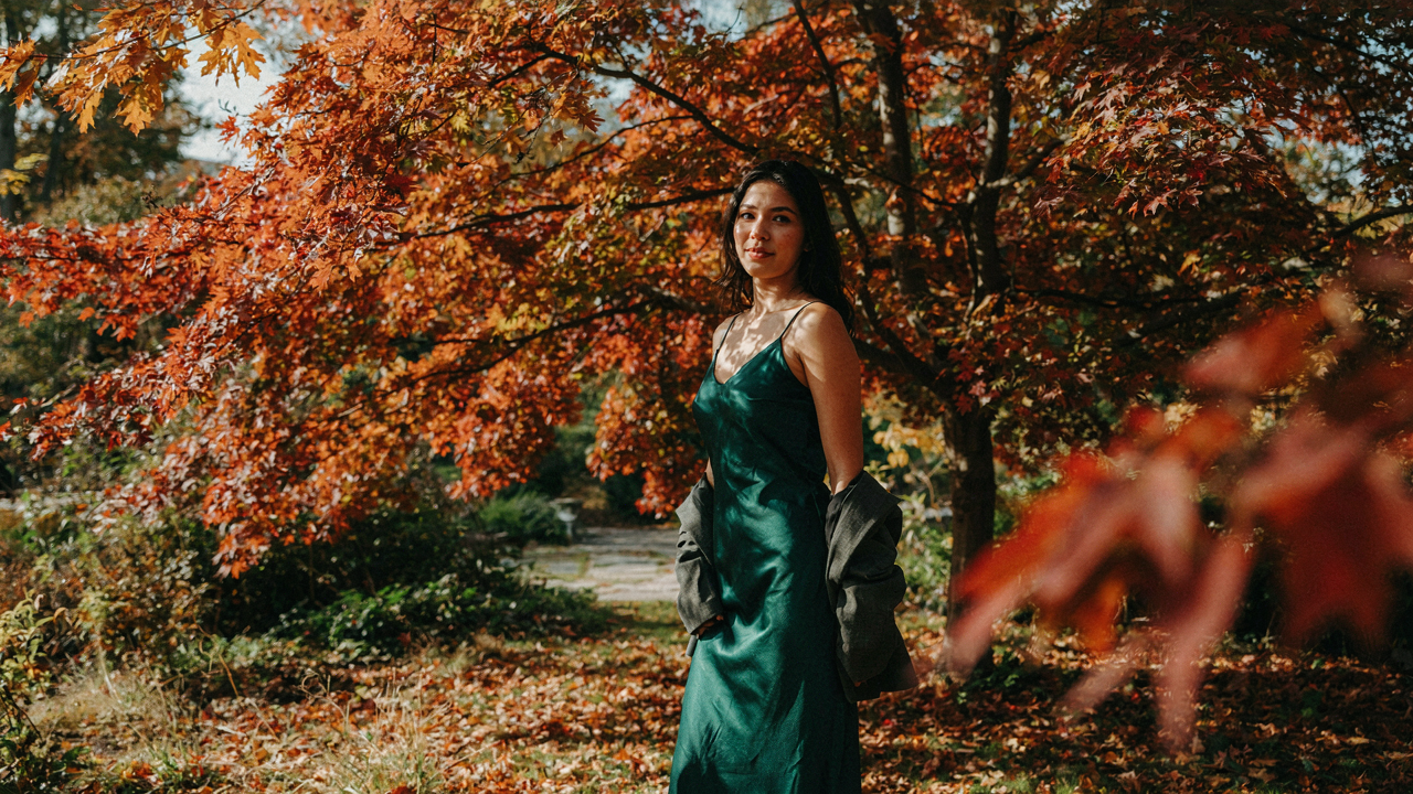 A woman in an emerald october wedding guest dress in a park with autumn leaves.