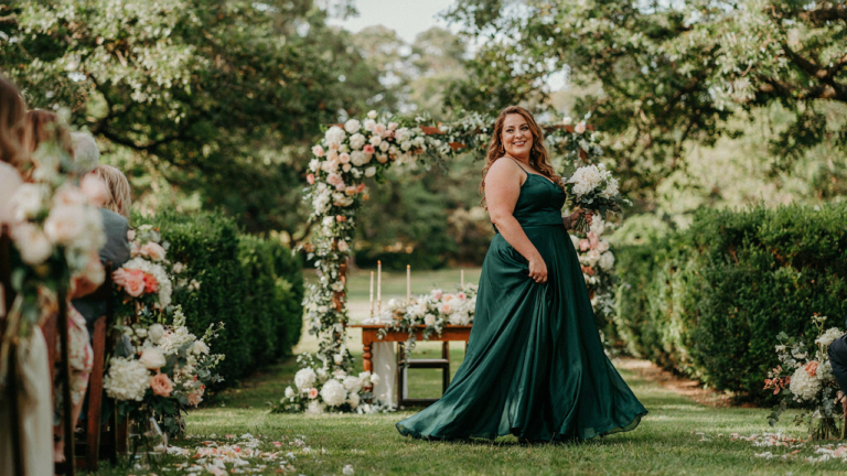 A smiling plus size wedding guest in a flowing dress walks confidently through a garden.