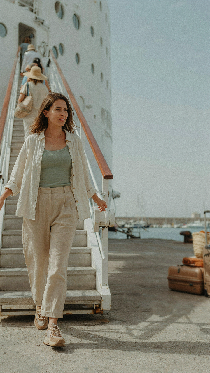 A woman in linen and walking shoes steps off a cruise ship at a sunny port.
