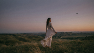 Woman in a long dress stands on a hill, overlooking a landscape at sunset.