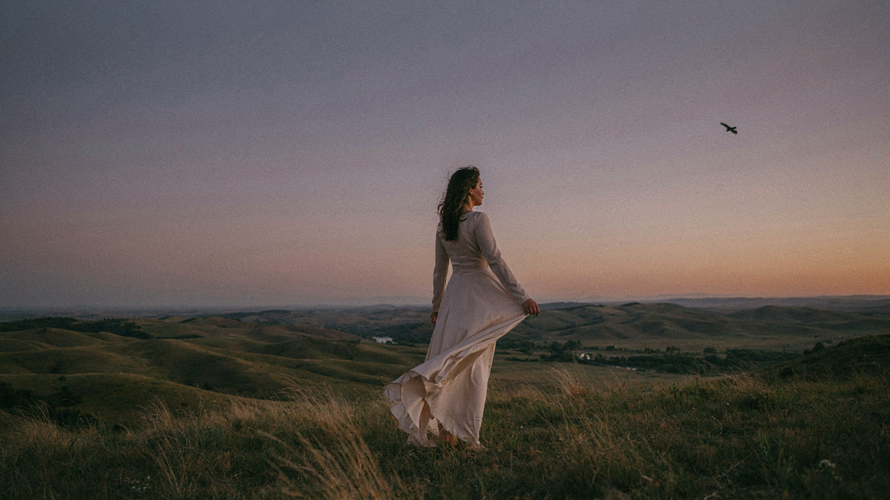 Woman in a long dress stands on a hill, overlooking a landscape at sunset.