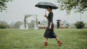 Woman in a dark midi dress and sturdy boots holds an umbrella at a rainy outdoor wedding.