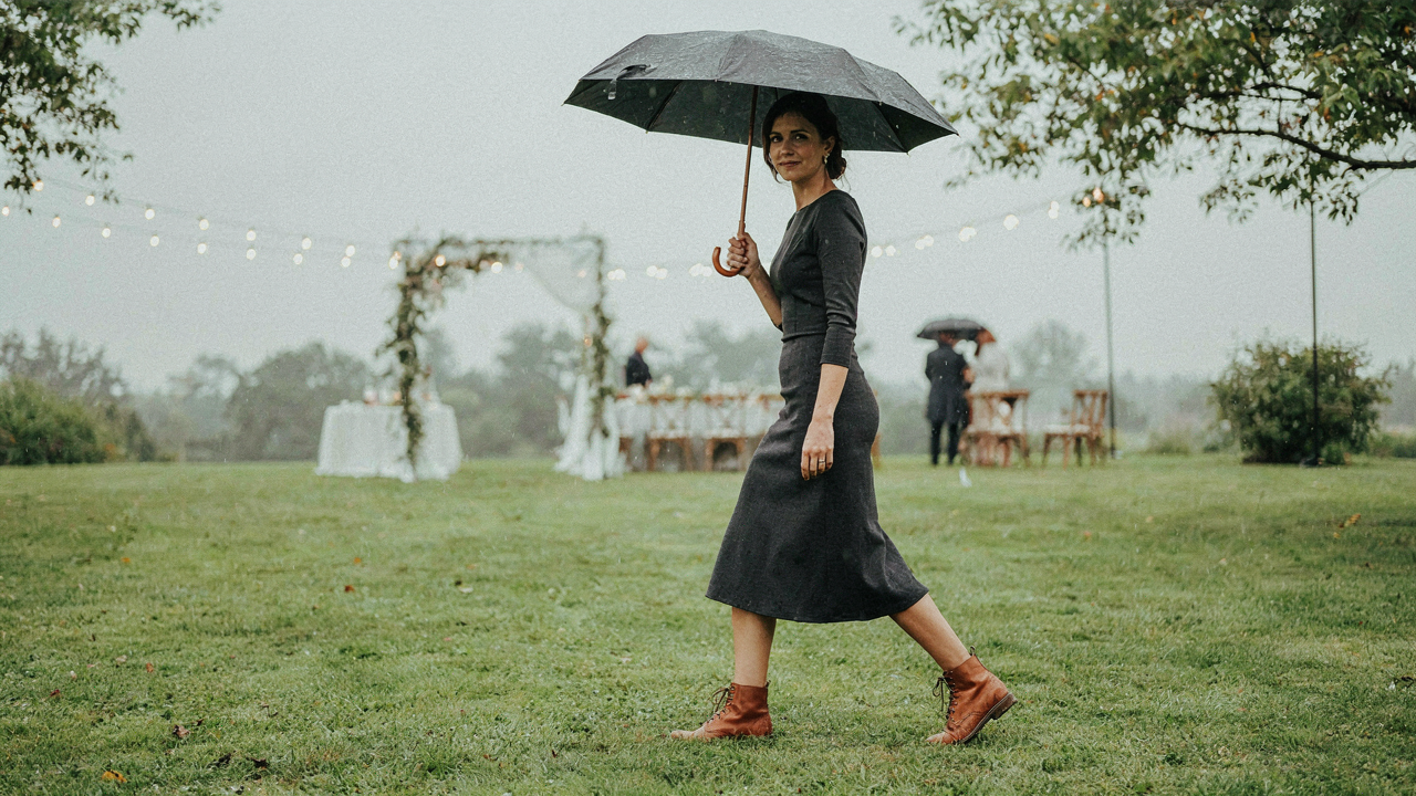 Woman in a dark midi dress and sturdy boots holds an umbrella at a rainy outdoor wedding.