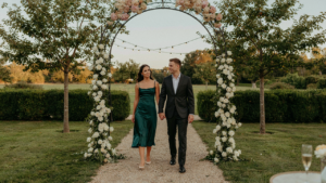 A smiling couple dressed in a cocktail dress and dark suit walks towards a garden party.