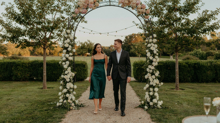 A smiling couple dressed in a cocktail dress and dark suit walks towards a garden party.