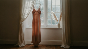 A beautiful blush silk dress with lace trim hangs by a window in soft morning light.