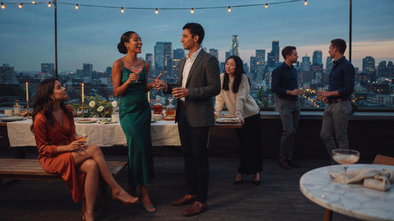 People chatting and laughing at a stylish rooftop cocktail party as the sun sets.