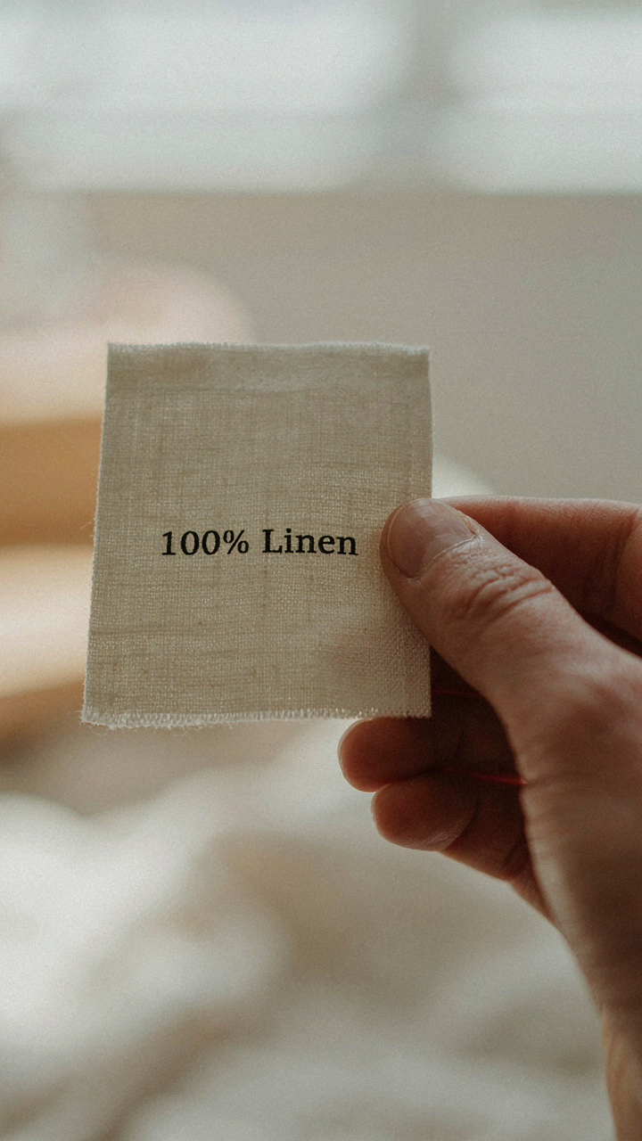 A hand holds a fabric tag clearly showing '100% Linen' on a rooftop cocktail party outfit.