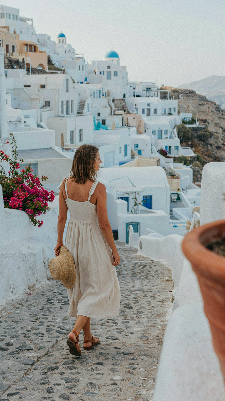 A woman in a linen dress and straw hat walks on a sunny Santorini path.