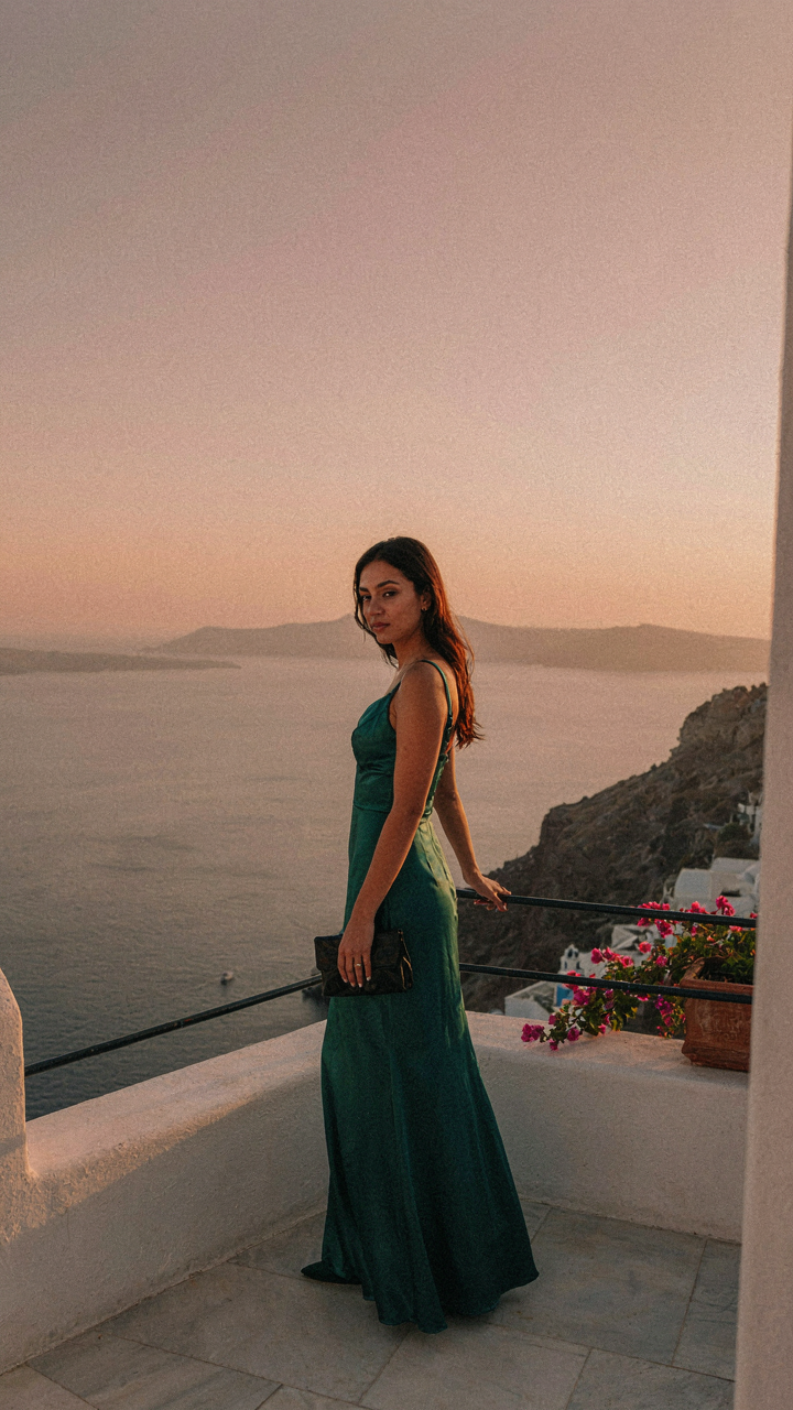 A woman in an elegant evening dress stands on a Santorini balcony at sunset.