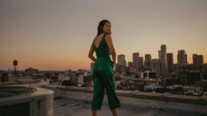 A woman in an emerald green satin jumpsuit stands on a city rooftop at sunset.