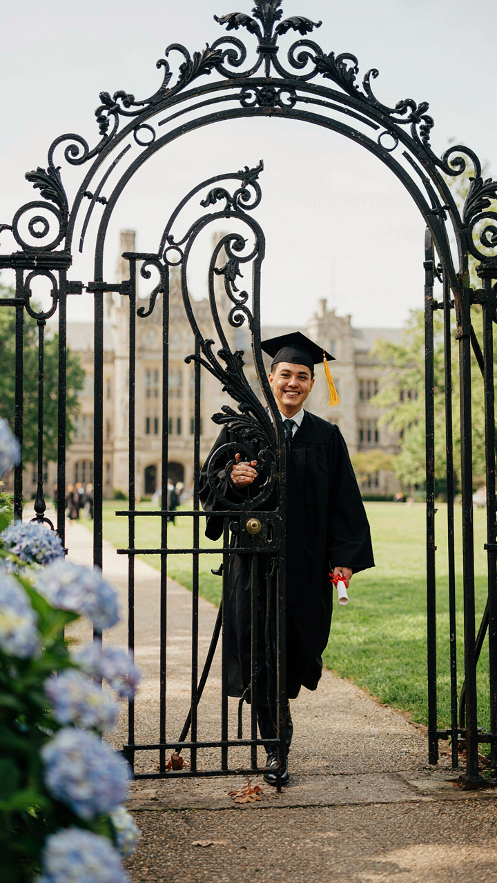 A person in a graduation gown walks through an ornate garden gate.