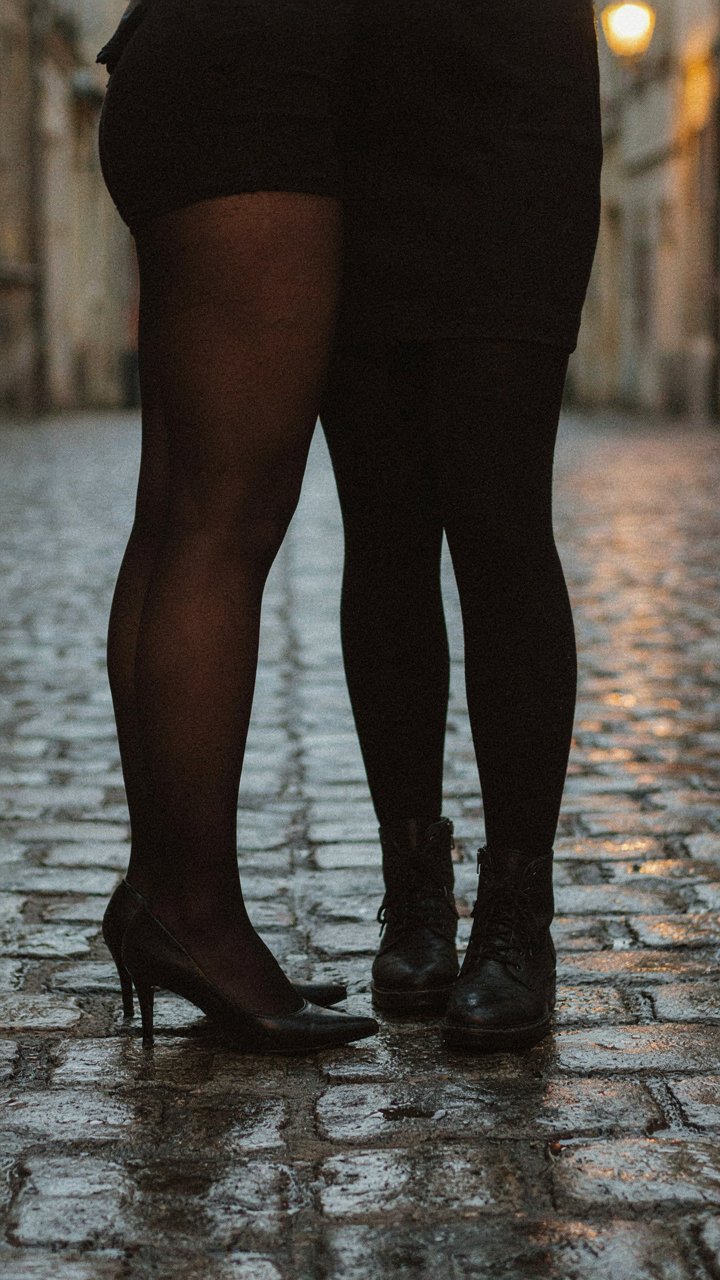 Two pairs of legs, one in sheer tights and one in opaque tights, stand on cobblestones.