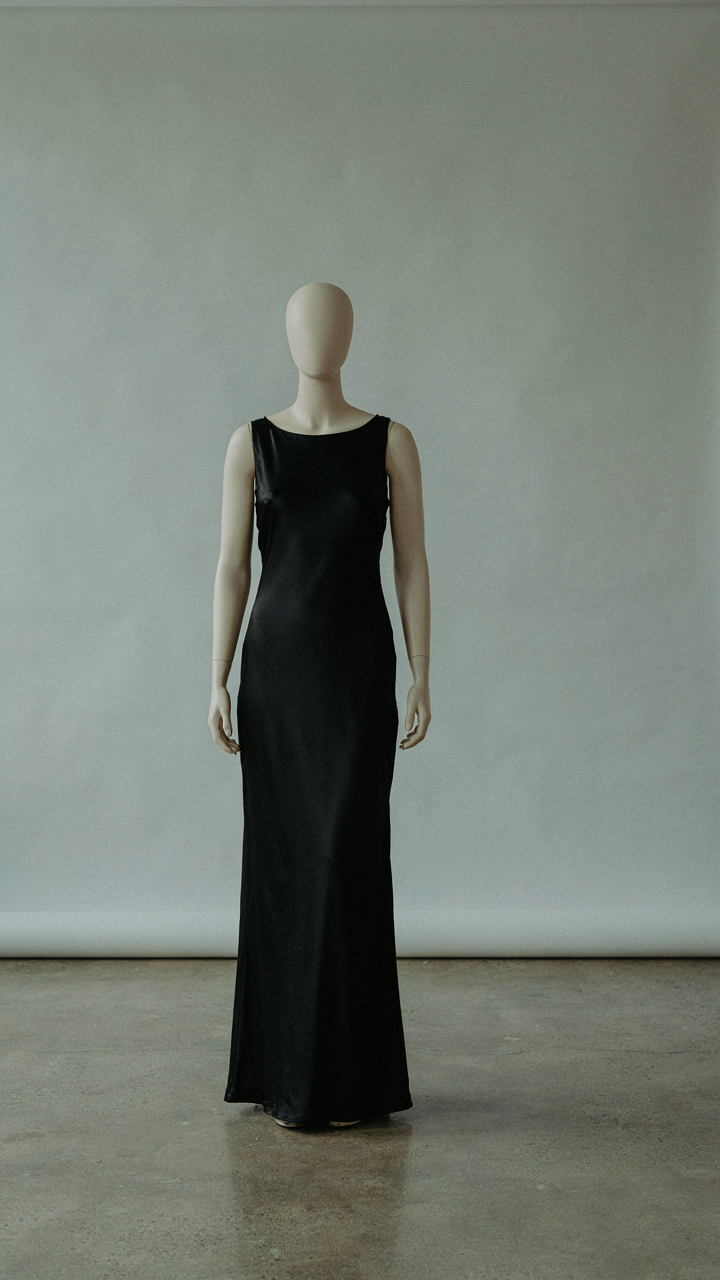 A minimalist black silk column gown stands on a mannequin in a studio.