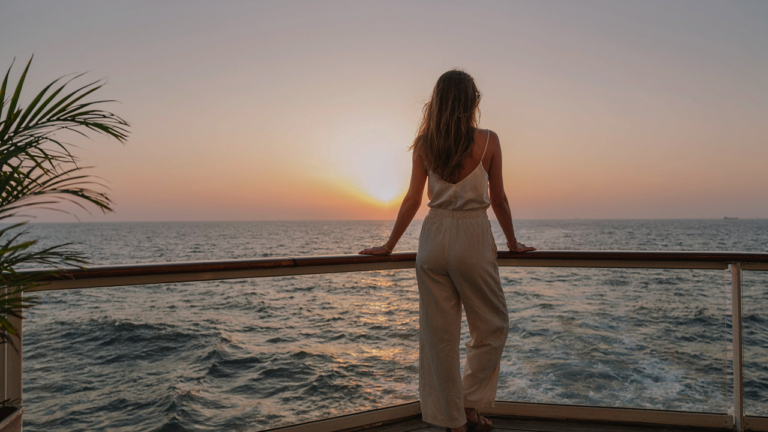 A woman in a stylish solo cruise outfit stands on a ship deck, gazing at the sunset.