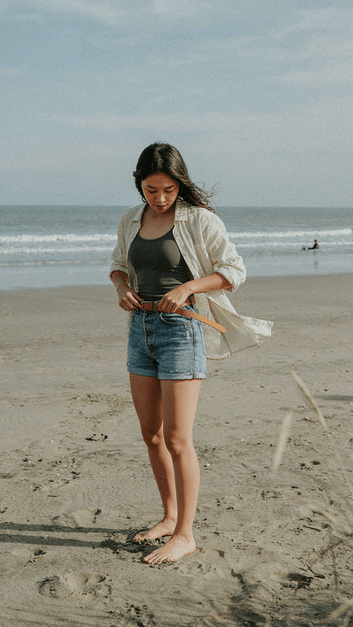 A person wears a linen overshirt and tank top, adjusting a belt on a windy beach.