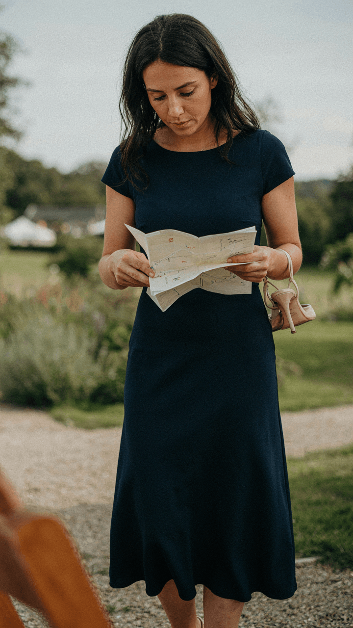 A woman in a blue midi dress looks at a map, worried about her heels on gravel.