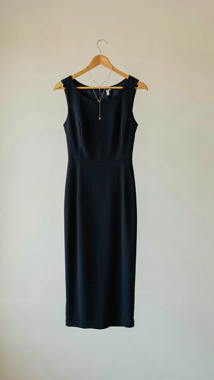 A navy sheath dress hangs on a hanger with a silver necklace.