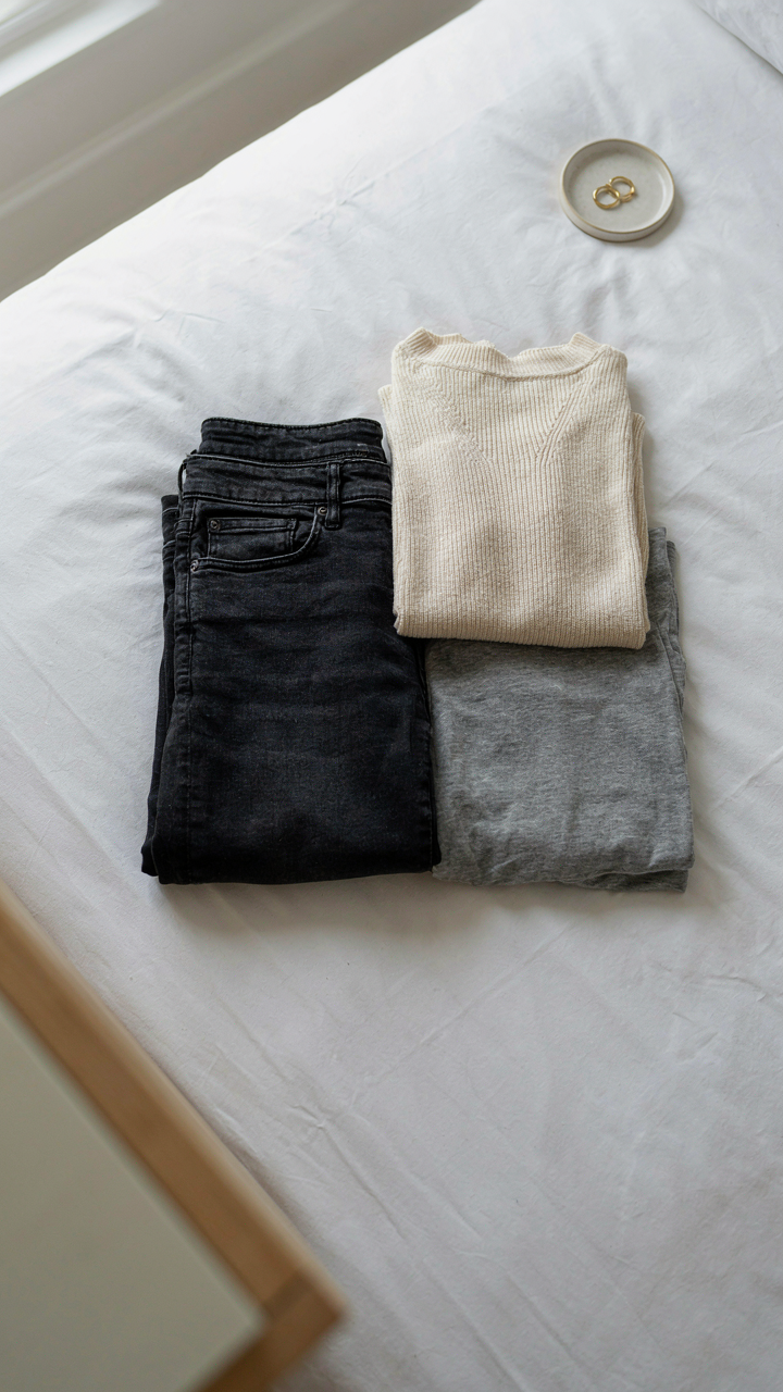 A pair of dark jeans and two different tops are neatly folded on a hotel bed.