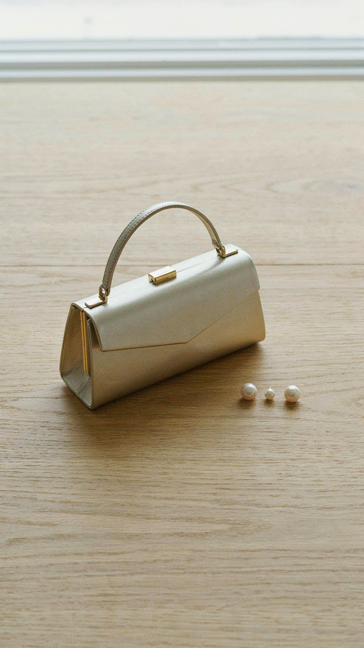 A structured metallic clutch bag and a single pearl earring on a wooden surface.