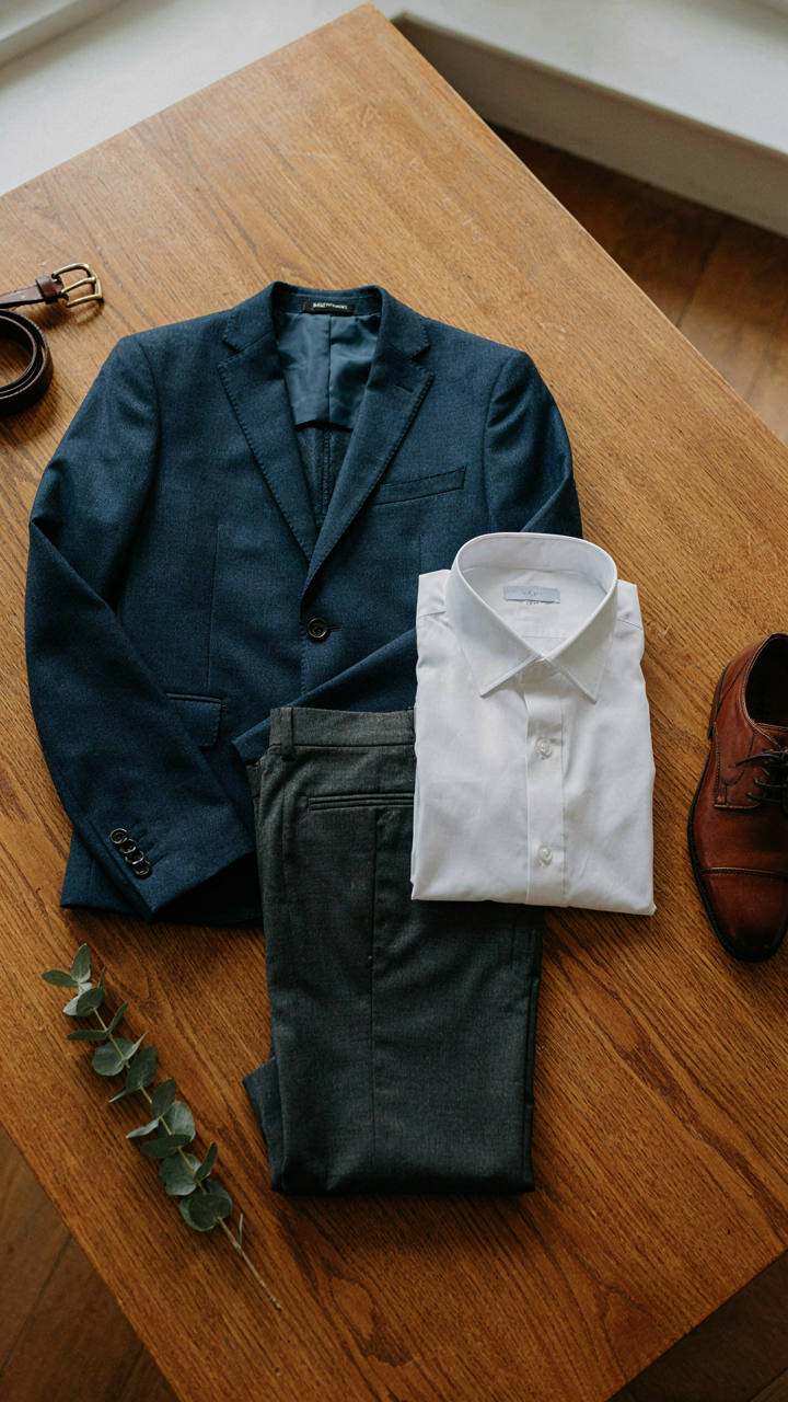 A perfectly pressed navy blazer, white dress shirt, and charcoal trousers on a wooden table.