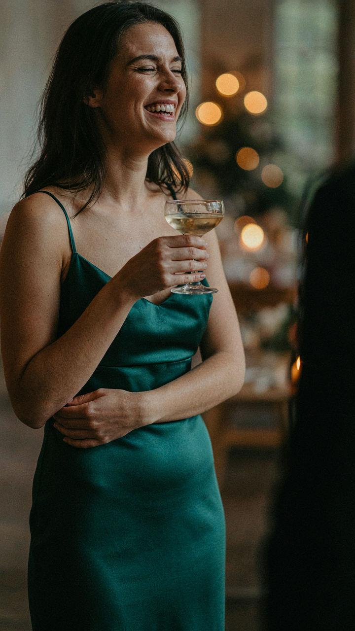 Woman in a cocktail dress laughs naturally during a conversation, holding a drink.