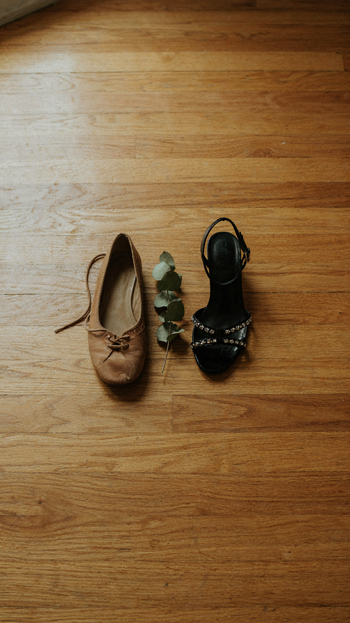 Worn ballet flats sit next to trendy embellished sandals on a wood floor.