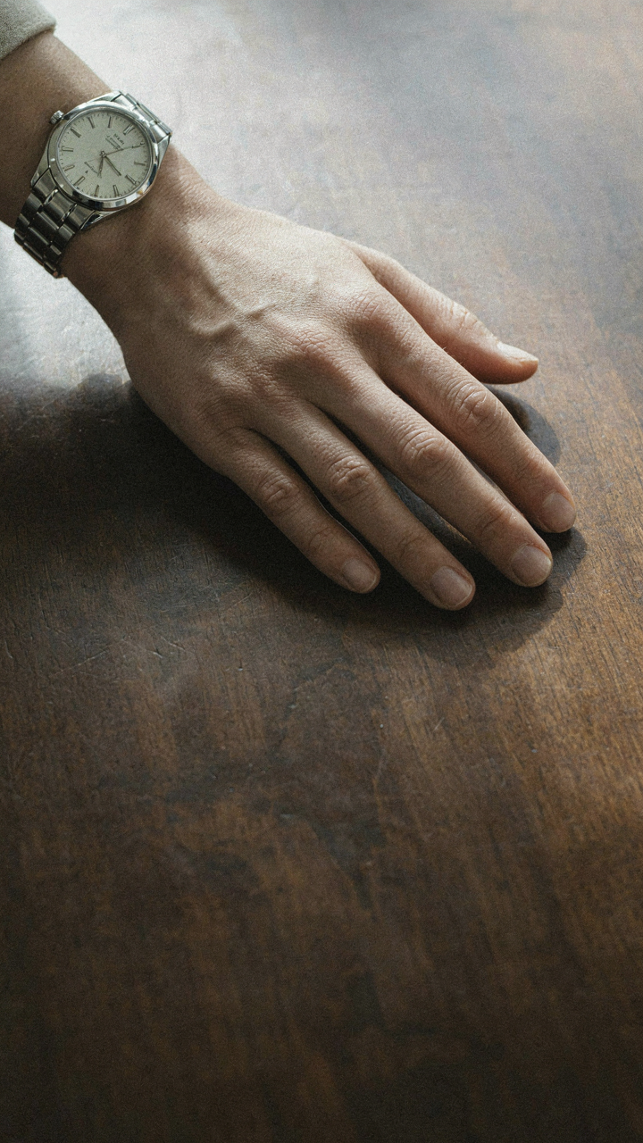 One elegant hand wears a classic metal watch, resting on a polished dark table.