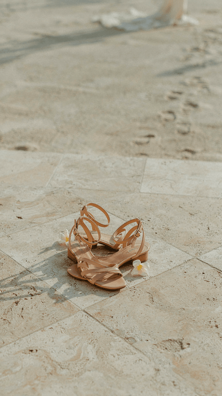 Elegant flat sandals sit on a tiled floor with sand nearby.