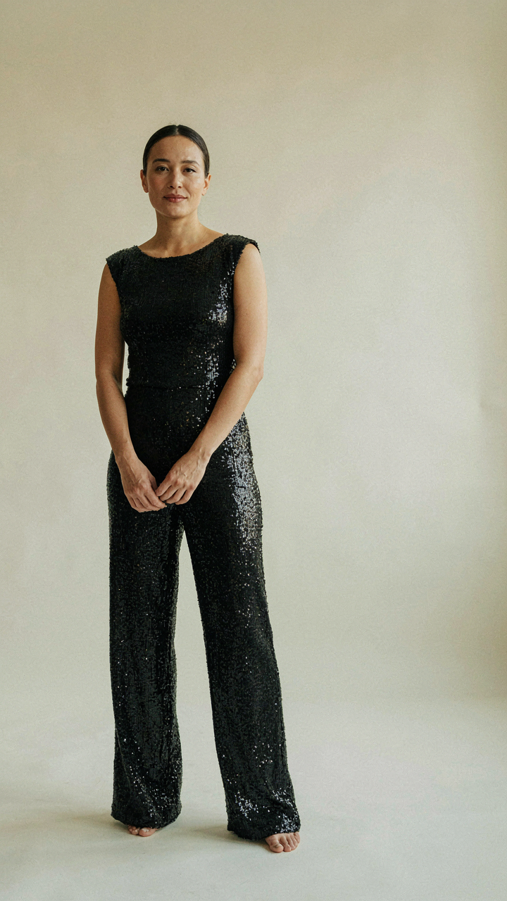 Woman in a black sequin jumpsuit standing still and looking confident.
