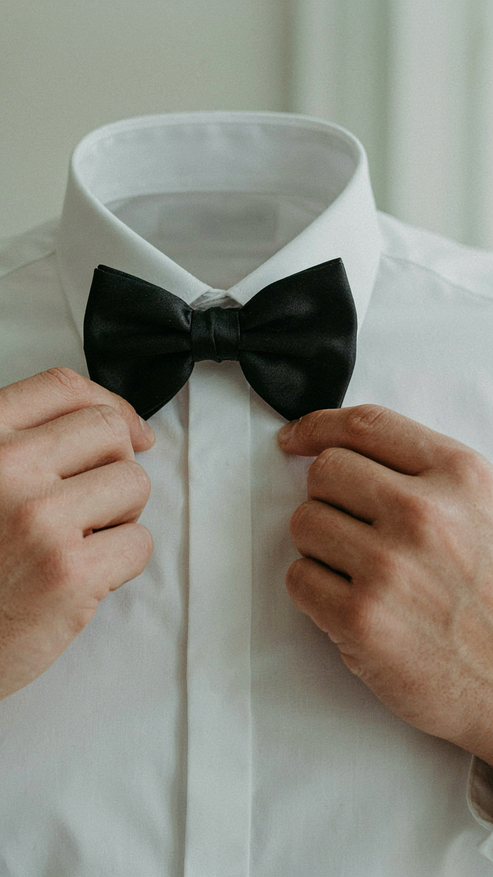 A man adjusts his black silk bow tie on a white dress shirt.