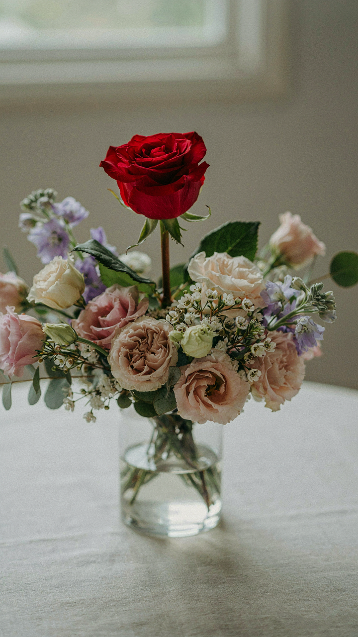 A vibrant red rose stands out among muted pastel flowers in a vase.