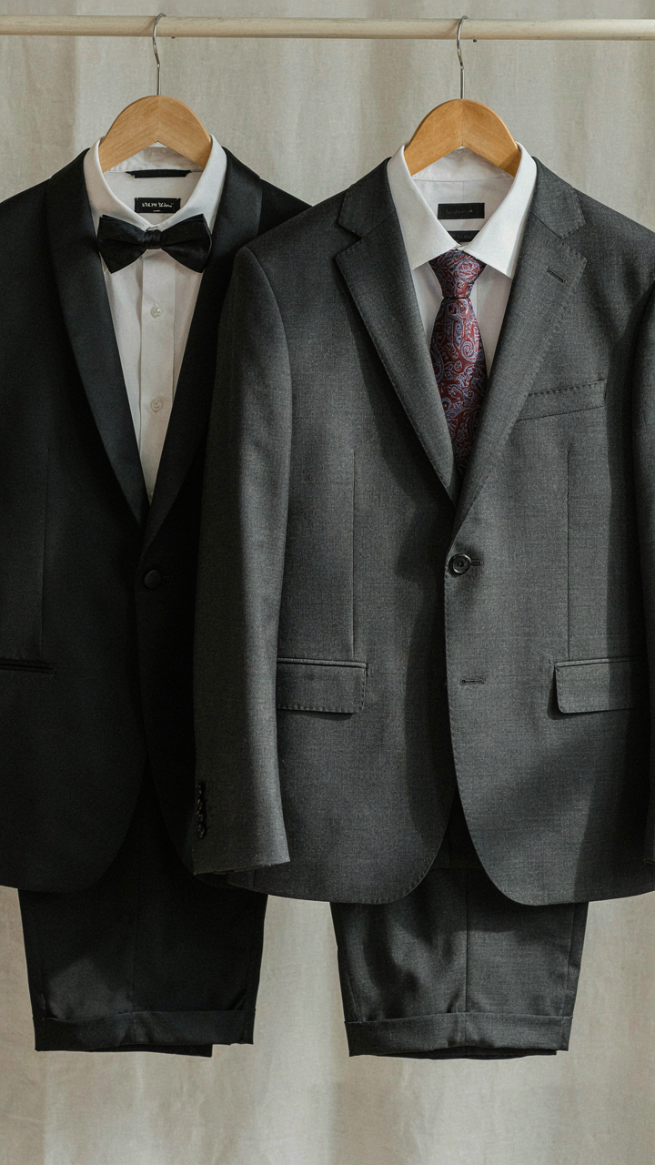 A tuxedo hangs next to a dark business suit on hangers, showing formal versus semi-formal.