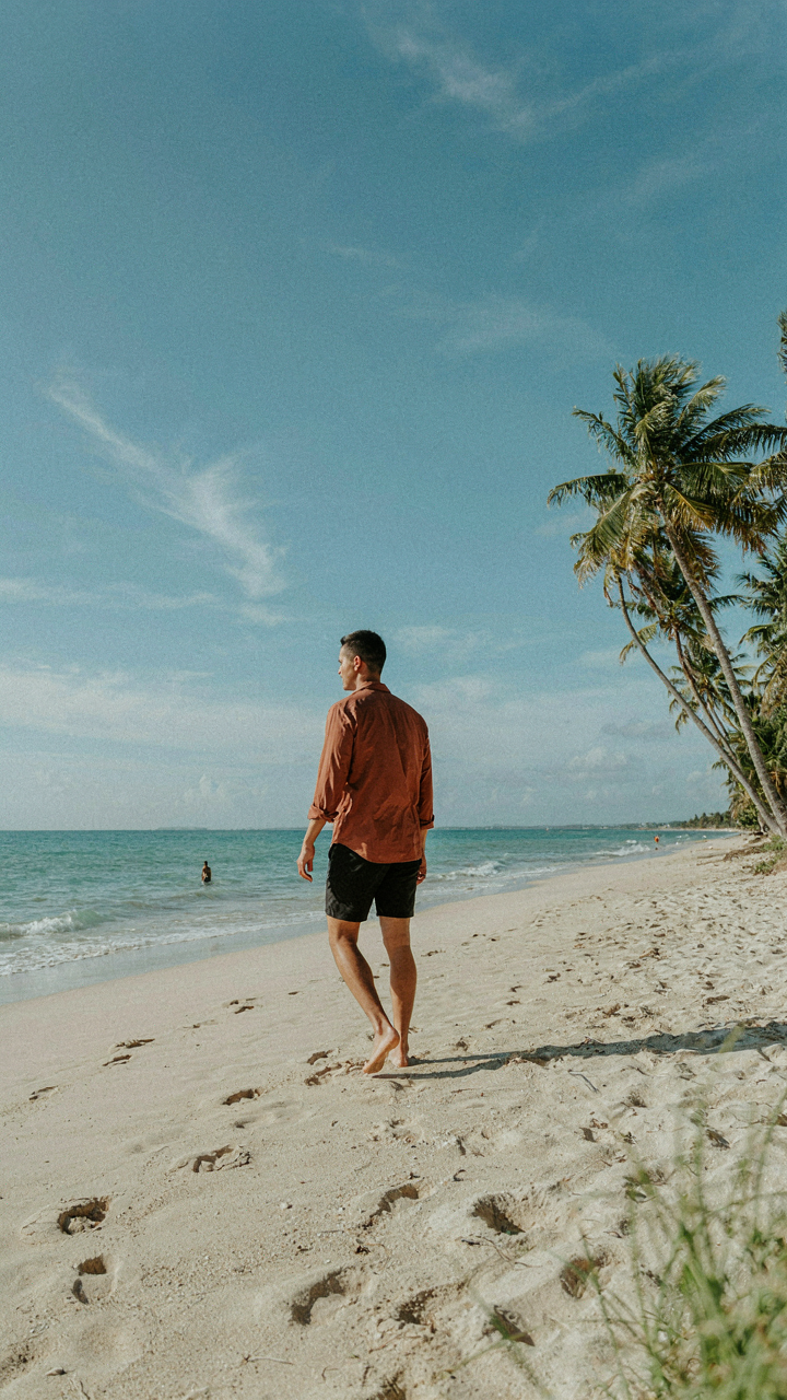 A person walking on a beach in a terracotta-colored shirt.