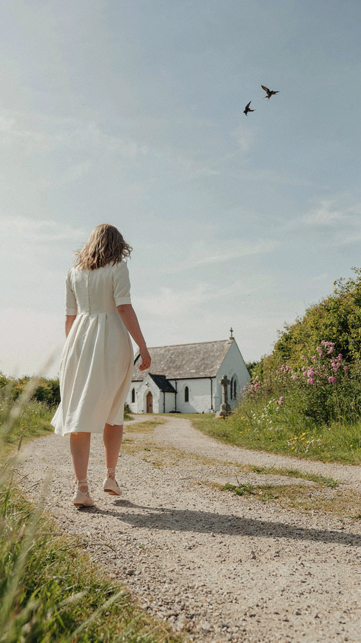 A woman in a cotton dress and ballet flats walks toward a country church.