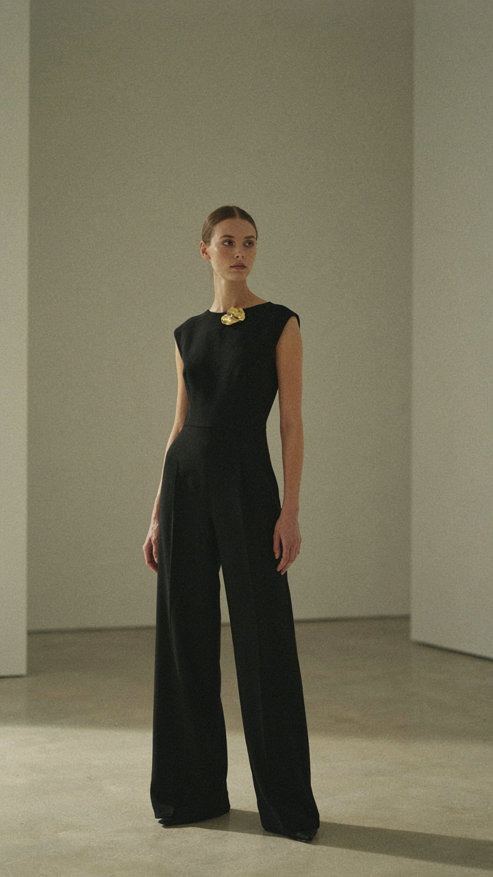 A person in a black structured jumpsuit with a single statement brooch.