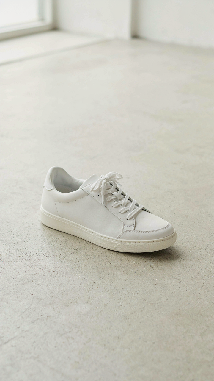 A simple, clean white leather sneaker sits on a light-colored floor.