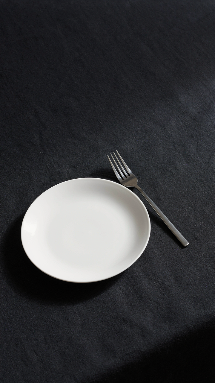 An elegant silver fork rests beside a white plate on a dark table.