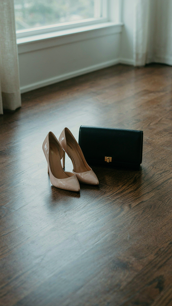 Nude stiletto pumps and a black clutch bag on a wooden floor.