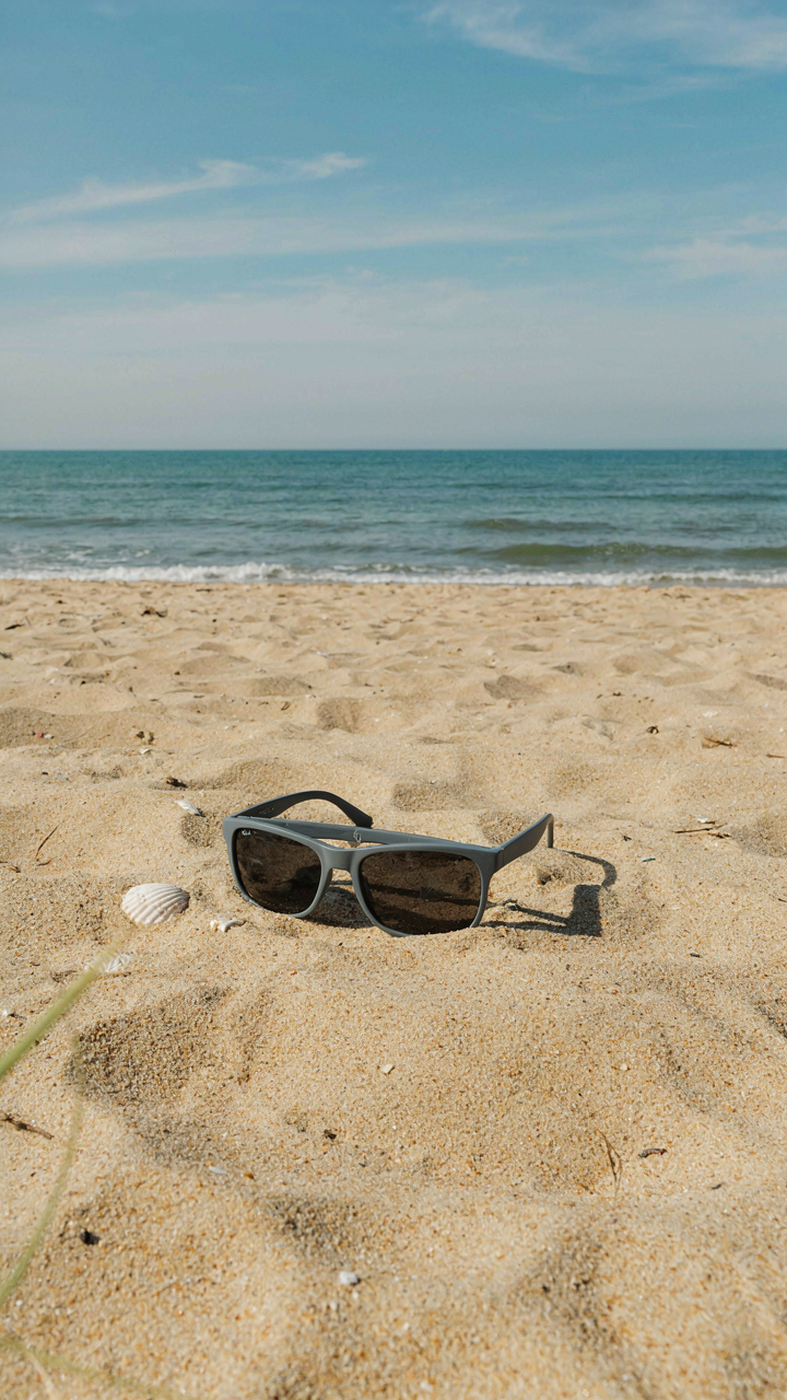 Gray polarized sunglasses rest on a sandy beach with the ocean in the background.