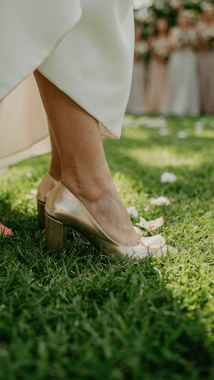 A woman's metallic block heels sink slightly into green grass.