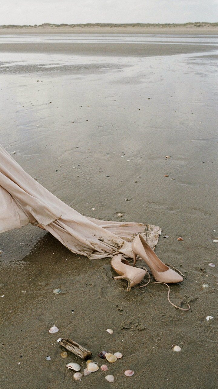 A ruined cocktail dress hem dragging through wet sand with sinking shoes.