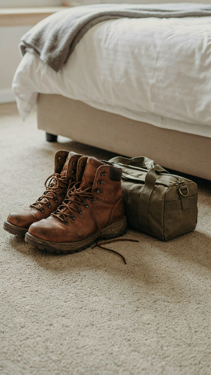 Large hiking boots sit unused on a hotel floor next to a small, packed travel bag.