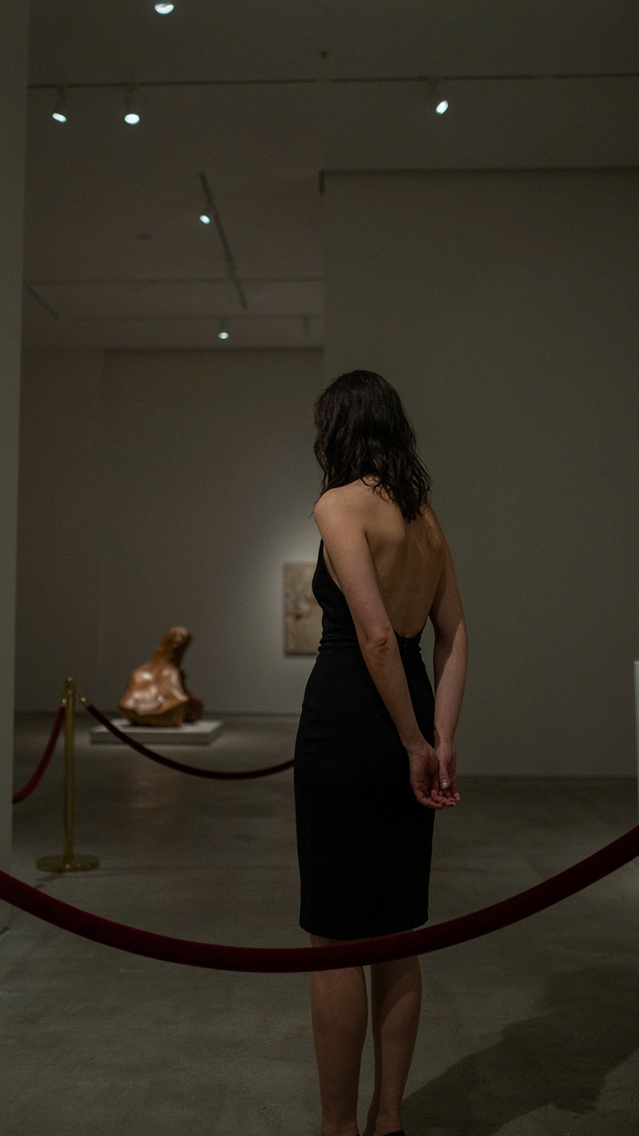 A woman in a backless cocktail dress looks at a covered sculpture in an art gallery.