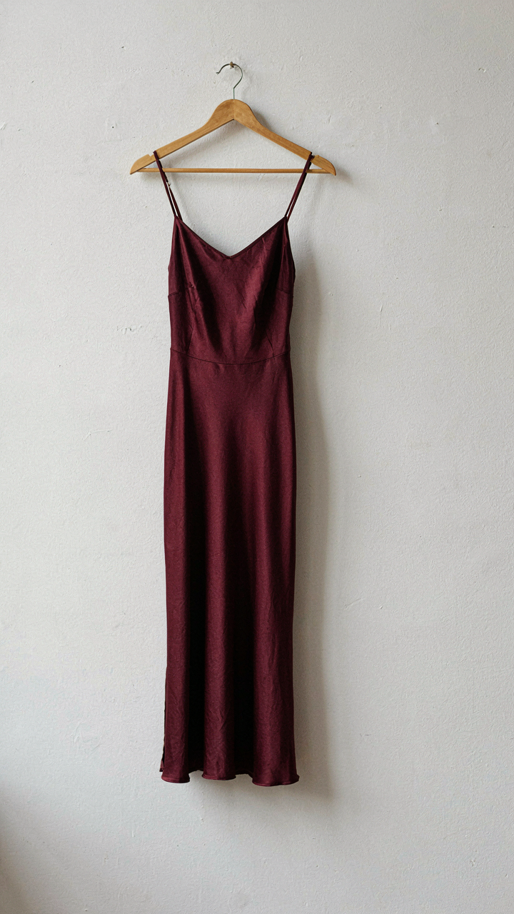 A simple burgundy silk slip dress hangs on a wooden hanger.