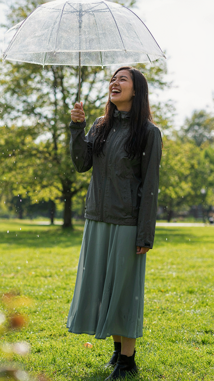 A woman under an umbrella, wearing a rain jacket and dress on a rainy day.
